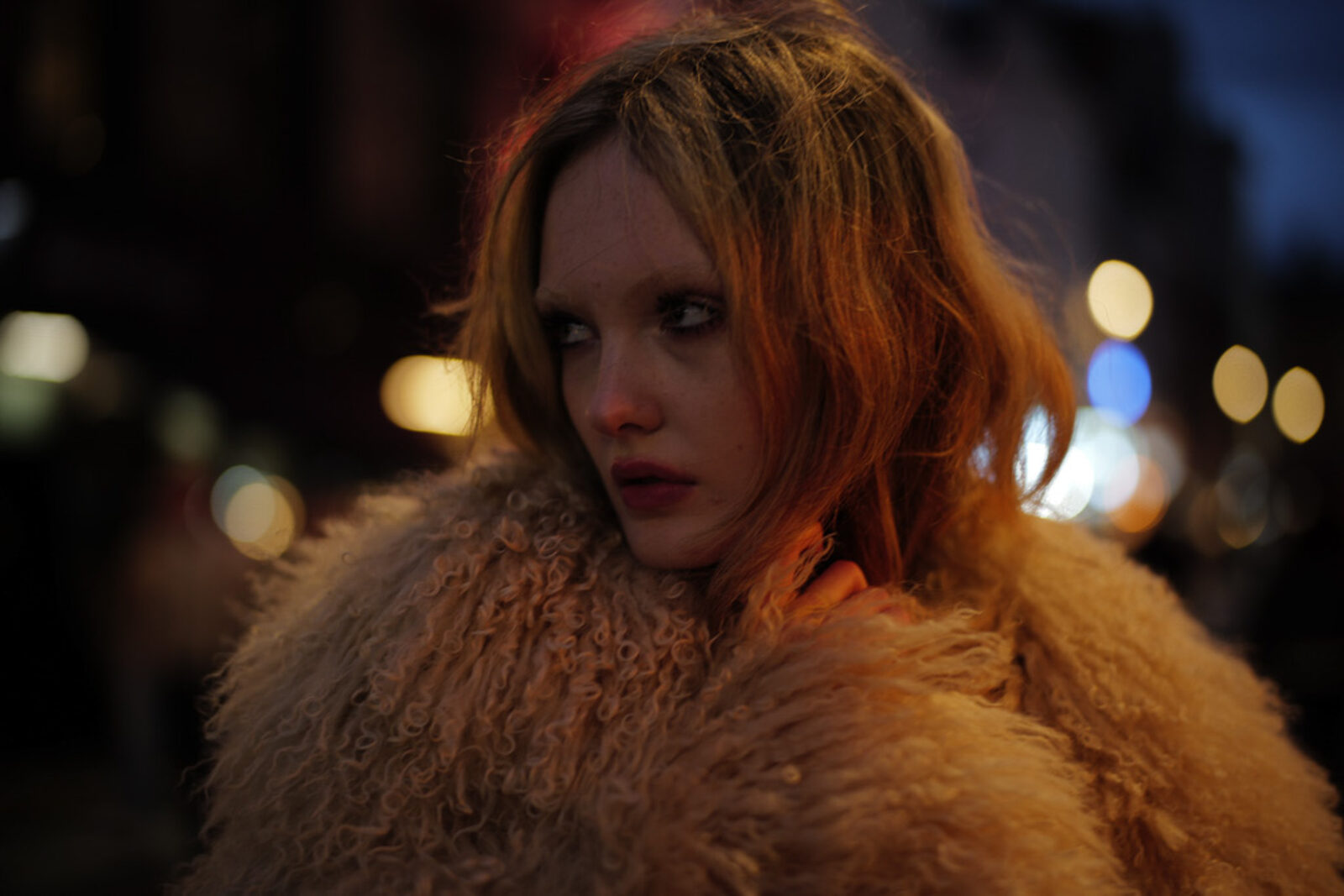 Night street portrait of a woman in a shaggy coat, city lights behind her
